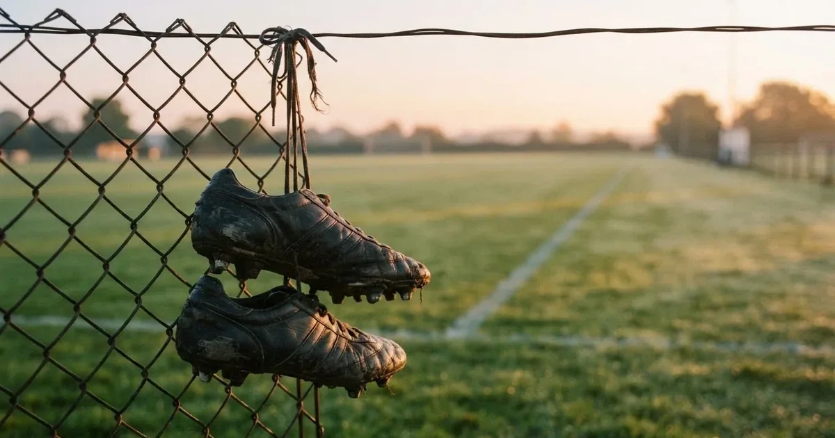 Znoszone buty piłkarskie zawieszone za sznurówki na drucianym płocie na skraju boiska treningowego o wschodzie słońca.